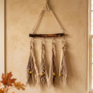 May include: A wall hanging featuring a dark brown horizontal wooden branch suspended by a thick cream-colored rope. Four bundles of dried beige and mauve pampas grass hang from the branch, each accented with a single small, round yellow flower.