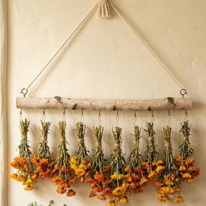 May include: A wall-hanging display featuring a horizontal birch branch suspended by a rope. Ten individual bundles of dried orange marigold flowers are attached to the branch with metal hooks, hanging downward in a uniform row.