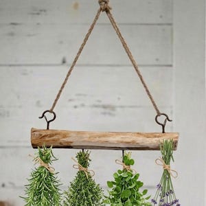 Op de afbeelding: Een horizontaal stuk natuurlijk lichtgekleurd hout opgehangen aan een dik touw van jute. Vier bundels verse groene kruiden en paarse lavendelbloemen zijn vastgebonden met touw en hangen aan metalen haken onderaan het hout.
