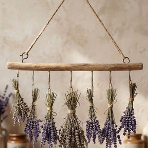Può includere: Un pezzo orizzontale di legno galleggiante di colore chiaro sospeso da una corda di juta intrecciata. Sette ganci metallici sono fissati al legno, ognuno dei quali sostiene un piccolo mazzetto legato di fiori di lavanda essiccati viola.