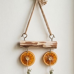 Puede incluir: Una decoración de pared que presenta una pieza de madera horizontal suspendida por una cuerda de yute gruesa. Dos rodajas de naranja secas están unidas a la madera, cada una con un pequeño manojo de hierbas verdes atadas con cordel.