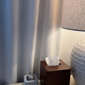 May include: A bedside scene featuring a gray speaker, a brown leather tissue box with white tissues, and a white lamp with a gray lampshade. The background includes a white curtain.