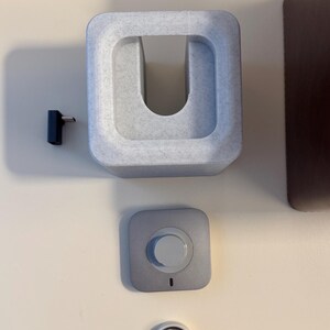 May include: A collection of tech accessories, including a light gray charging dock, a small silver-colored button, a black pin, and a silver ring. The charging dock has a U-shaped opening. The button has a white center and a small black indicator light.