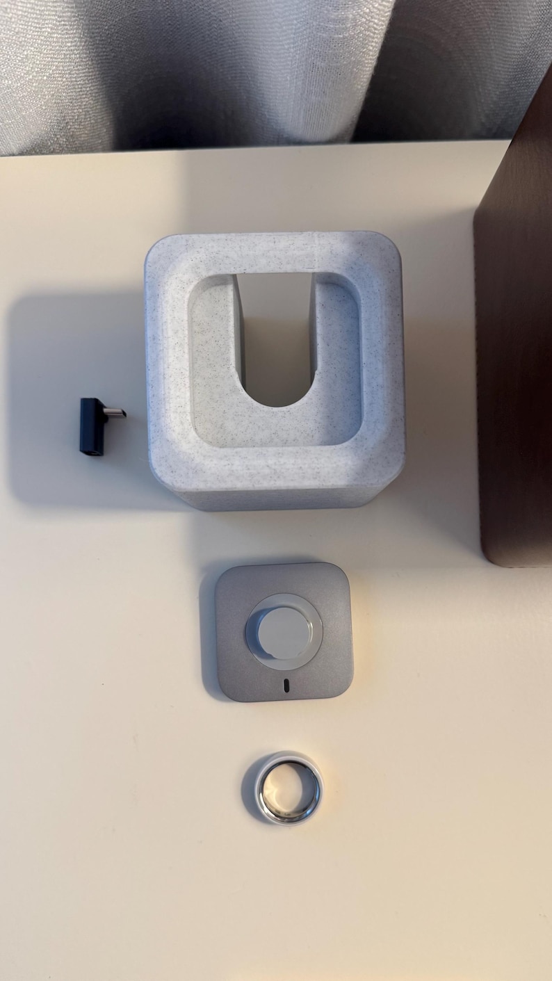 May include: A collection of tech accessories, including a light gray charging dock, a small silver-colored button, a black pin, and a silver ring. The charging dock has a U-shaped opening. The button has a white center and a small black indicator light.