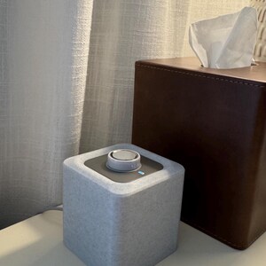 May include: A light gray, cube-shaped speaker with a silver control dial on top. A brown tissue box is behind the speaker. The speaker is on a white surface, with a white curtain in the background.