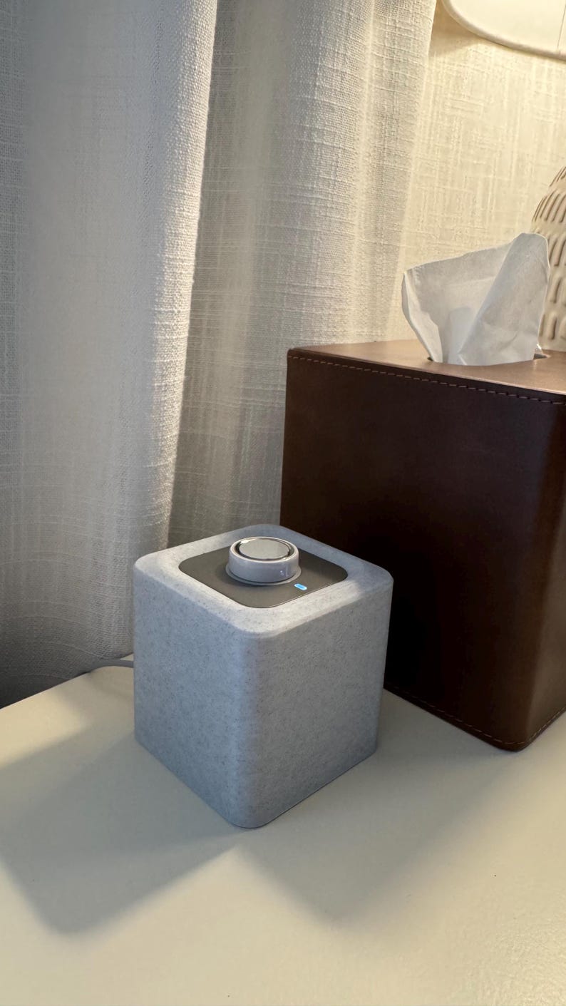 May include: A light gray, cube-shaped speaker with a silver control dial on top. A brown tissue box is behind the speaker. The speaker is on a white surface, with a white curtain in the background.