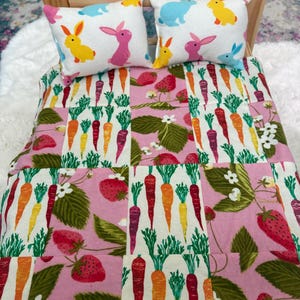 May include: A doll bed with two white pillows featuring colorful bunnies and a pink, white, and green quilt. The quilt has a pattern of carrots and strawberries.