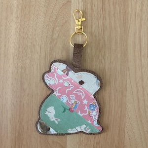 May include: A handmade bunny-shaped keychain with a gold-tone clasp. The bunny is made of patchwork fabric in pink, green, and white with a brown felt backing. The fabric features patterns of rabbits, moons, and stars.