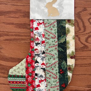 May include: A handmade Christmas stocking with a variety of festive patterns. The top is a light gray with a gold bunny silhouette. The body features stripes of snowmen, candy canes, holly, and pine boughs. A green ribbon loop is attached.
