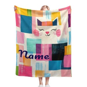 May include: A colourful blanket featuring a white cat face and the word "Name" in blue. The blanket has a geometric pattern of squares and rectangles in shades of pink, blue, yellow, green, and purple.