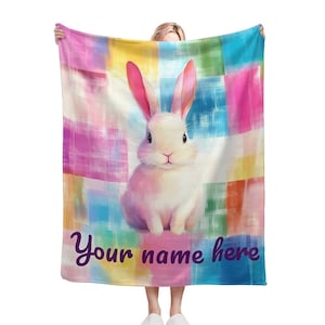 May include: A colourful blanket featuring a white bunny rabbit in the centre. The blanket has a checkered pattern with pink, blue, yellow and green colours. The text "Your name here" is printed at the bottom of the blanket.