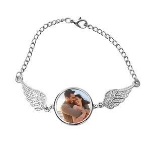 Puede incluir: Pulsera plateada con un colgante redondo que muestra la foto de una pareja. La pulsera tiene eslabones de cadena finos y dijes decorativos de alas. Un accesorio romántico y personalizado.