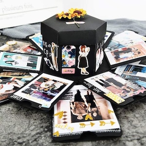 Puede incluir: Una caja de explosión negra con múltiples compartimentos, cada uno con una foto. La caja está decorada con flores, pegatinas y las palabras "ME TOO" y "TODAY".