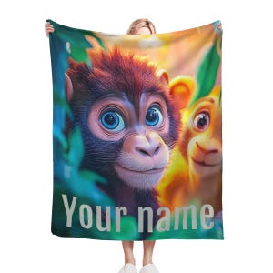 Könnte beinhalten: Eine bunte Fleece-Decke mit einer Nahaufnahme von zwei Cartoon-Affen mit großen blauen Augen. Der Text "Your name" ist unten auf der Decke aufgedruckt.