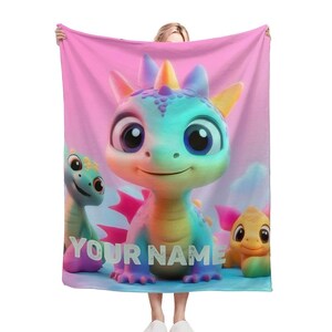 Puede incluir: Una manta de forro polar rosa con un dragón colorido y dos dragones más pequeños. El texto "YOUR NAME" está impreso en la manta.