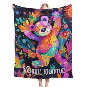 Pode incluir: Uma manta colorida com um design de urso brilhante e multicolorido. O urso está rodeado de flores e folhas. O texto "Your name" está impresso na parte inferior da manta.