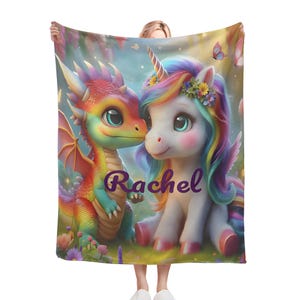 Puede incluir: Una manta colorida con un dragón y un unicornio. El dragón tiene escamas de arcoíris, y el unicornio tiene una melena de arcoíris y una corona de flores. El nombre "Rachel" está escrito en púrpura en la manta.