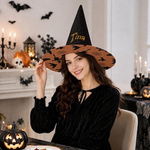 May include: A pointed witch hat featuring a black cone with the name Tina embroidered in gold thread. The wide brim is tan with a repeating black pattern of bats and spiderwebs. The hat has a structured shape and a textured fabric finish.