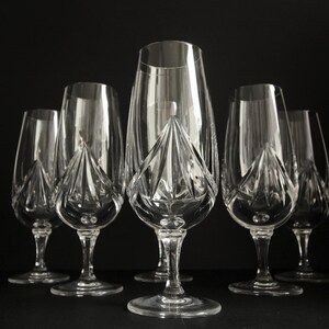 Könnte beinhalten: Ein Set aus sechs klaren Kristall-Weingläsern. Jedes Glas hat ein einzigartiges, facettiertes Design am Boden der Kelchschale. Die Gläser sind vor einem dunklen Hintergrund angeordnet, was ihre elegante Form und Klarheit hervorhebt.