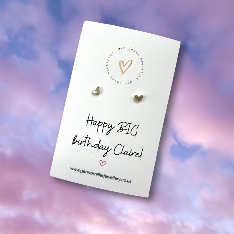 Pode incluir: Brincos de bot&atilde;o dourados em forma de cora&ccedil;&atilde;o em um cart&atilde;o branco com o texto "Happy BIG birthday Claire!" e o endere&ccedil;o do site gennamillerjewellery.co.uk. O cart&atilde;o tem um logotipo com um design de cora&ccedil;&atilde;o. O fundo &eacute; um c&eacute;u suave roxo e rosa.