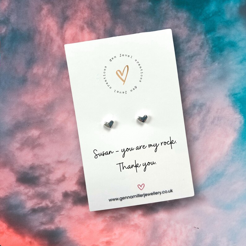 Pode incluir: Um cart&atilde;o branco com brincos de cora&ccedil;&atilde;o prateados. O cart&atilde;o tem o texto "Susan - you are my rock. Thank you." e um endere&ccedil;o web. O fundo &eacute; uma mistura de rosa e azul.