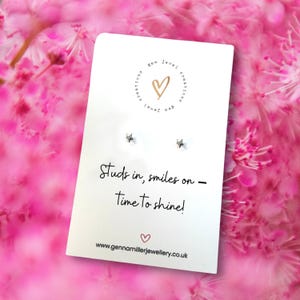 Pode incluir: Um cartão branco com o texto "Studs in, smiles on - Time to shine!" e um endereço de site, com dois pequenos brincos em forma de estrela. O cartão tem um logotipo com um coração. O fundo são flores cor-de-rosa.
