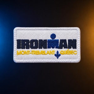 May include: A rectangular embroidered patch with a white background and a stitched border. The text IRONMAN is in dark blue, with a stylized blue figure above the letter M. Below, the text MONT-TREMBLANT QUÉBEC is in yellow with a blue fleur-de-lis.