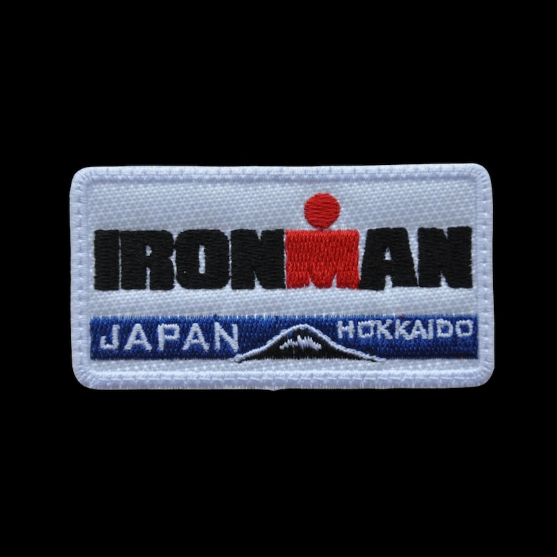 May include: Embroidered patch with the text "IRONMAN" in black and red, with a red figure above the word. Below, the text "JAPAN" and "HOKKAIDO" flank a mountain graphic, all on a white background.