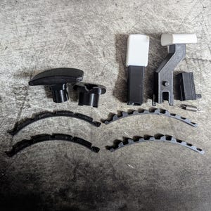 Könnte beinhalten: Eine Ansammlung von schwarz-weißen 3D-gedruckten Teilen, darunter gebogene Elemente, Knöpfe und rechteckige Komponenten. Die Teile sind auf einer strukturierten grauen Oberfläche angeordnet und zeigen verschiedene Formen und Größen.