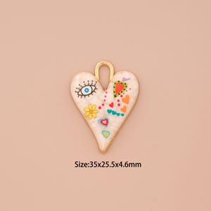Könnte beinhalten: Goldfarbener herzförmiger Anhänger mit weißer Emaille-Oberfläche. Der Anhänger ist mit bunten, bemalten Details verziert, darunter ein blaues Auge, eine gelbe Blume und rote, rosa und grüne Herzen. Der Anhänger misst 3,5 x 2,5 x 4,6 cm.