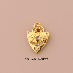 Puede incluir: Colgante dorado en forma de escudo, adornado con cristales transparentes y una piedra central en forma de lágrima. El colgante tiene un lazo en la parte superior para colgar y el texto "Size: 16.1x11x2.8mm".