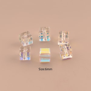 May include: Six iridescent, clear cube beads, each measuring 6mm, are scattered on a light peach surface. The beads reflect a rainbow of colors, with visible internal structures. The text "Size: 6mm" is displayed below the beads.