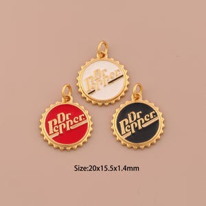 Puede incluir: Tres colgantes dorados con forma de tapones de botella, cada uno con un esquema de color diferente: rojo, blanco y negro. El logotipo de Dr Pepper se muestra en cada colgante. Tamaño: 20x15,5x1,4 mm.