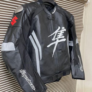 May include: Black leather motorcycle jacket with white and gray accents. The jacket has "Hayabusa" written on the left sleeve and "隼" on the back. The jacket has a silver Suzuki logo on the left chest.