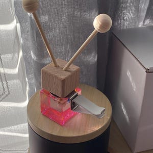Puede incluir: Un frasco cuadrado de vidrio para ambientador de coche lleno de líquido rosa brillante, rematado con un cubo de madera clara y dos palitos de madera con extremos esféricos. Un clip de plástico blanco está unido al lado del recipiente.