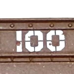 May include: Close-up of a weathered, brown metal surface with the white stenciled text "G. 100 LT." Rivets and metal brackets are visible along the top and sides of the surface. The image suggests a vintage industrial or transportation theme.