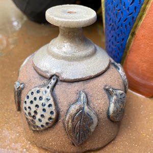 Op de afbeelding: Een kleine, keramische pot met een lichtbruine glazuur en een lichtgrijze bovenkant. De pot is versierd met vier bladeren, elk met een ander ontwerp. De pot heeft een klein gaatje in de bovenkant.