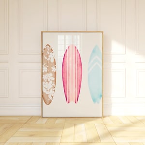 May include: A framed print of three watercolor surfboards. The surfboards are in different colors and patterns. The left surfboard is brown with white flowers, the middle surfboard is pink with white stripes, and the right surfboard is blue with white chevron stripes.