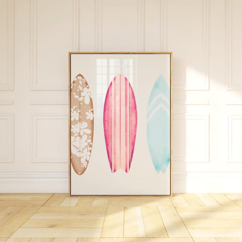 May include: A framed print of three watercolor surfboards. The surfboards are in different colors and patterns. The left surfboard is brown with white flowers, the middle surfboard is pink with white stripes, and the right surfboard is blue with white chevron stripes.