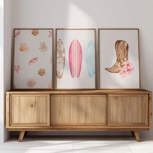 May include: Three framed prints with watercolor illustrations. The left print features a white background with brown seashells. The center print features three surfboards in pink, blue, and white. The right print features a brown cowboy boot with pink flowers.