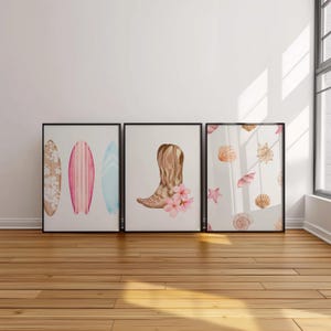 May include: Three framed watercolor prints. The first print features two surfboards with pink, blue, and white designs. The second print features a brown cowboy boot with pink flowers. The third print features a white background with a pattern of seashells and a starfish.