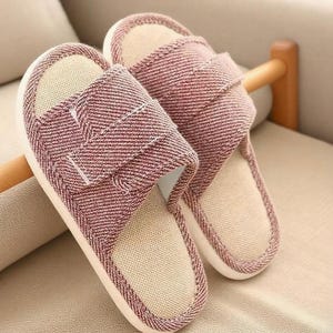 May include: A pair of open-toe house slippers. The slippers have a red and white herringbone pattern on the straps and sides, with a beige interior. The soles are white. The slippers are resting on a beige surface.