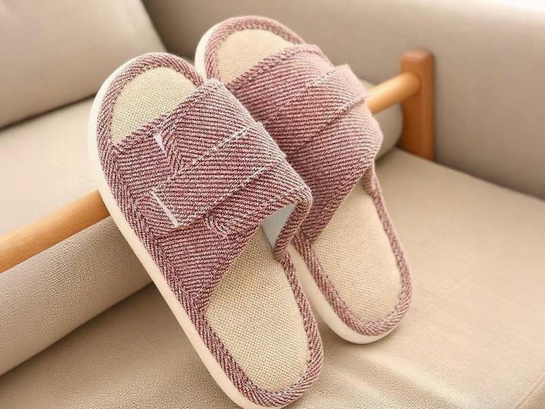 May include: A pair of open-toe house slippers. The slippers have a red and white herringbone pattern on the straps and sides, with a beige interior. The soles are white. The slippers are resting on a beige surface.