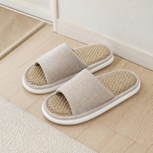 May include: A pair of open-toe slide slippers featuring a beige fabric upper with a diagonal weave pattern. The footbed is constructed from a textured, woven natural fiber material, and the slippers are finished with a smooth white rubber sole.