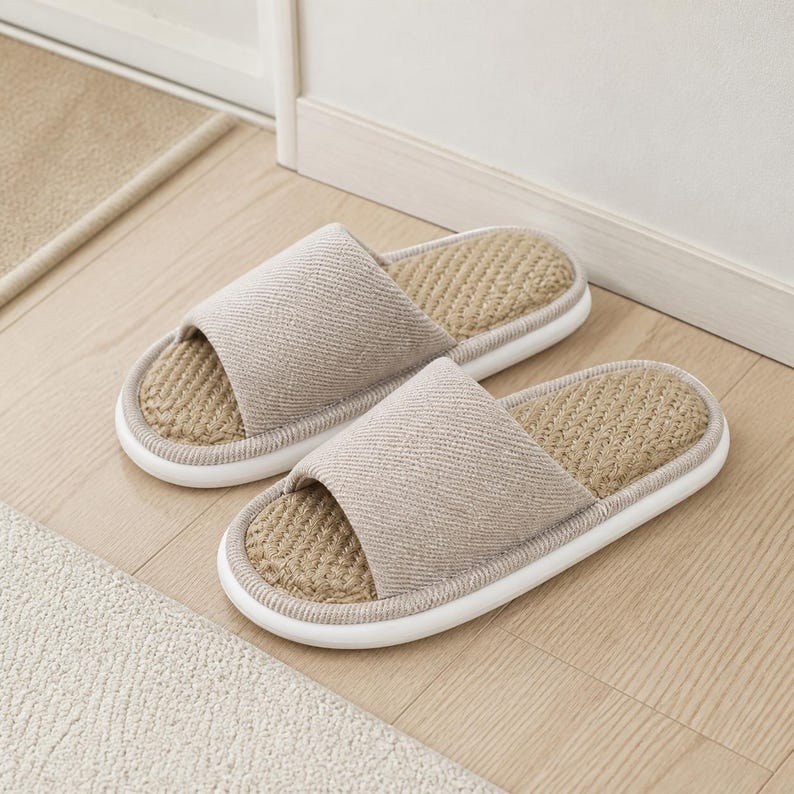 May include: A pair of open-toe slide slippers featuring a beige fabric upper with a diagonal weave pattern. The footbed is constructed from a textured, woven natural fiber material, and the slippers are finished with a smooth white rubber sole.