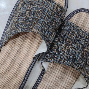 May include: A pair of open-toe slippers featuring a woven tan straw-like footbed and a wide upper strap made of a textured fabric in shades of dark blue, brown, and light blue. The edges are finished with dark brown piping and white stitching.