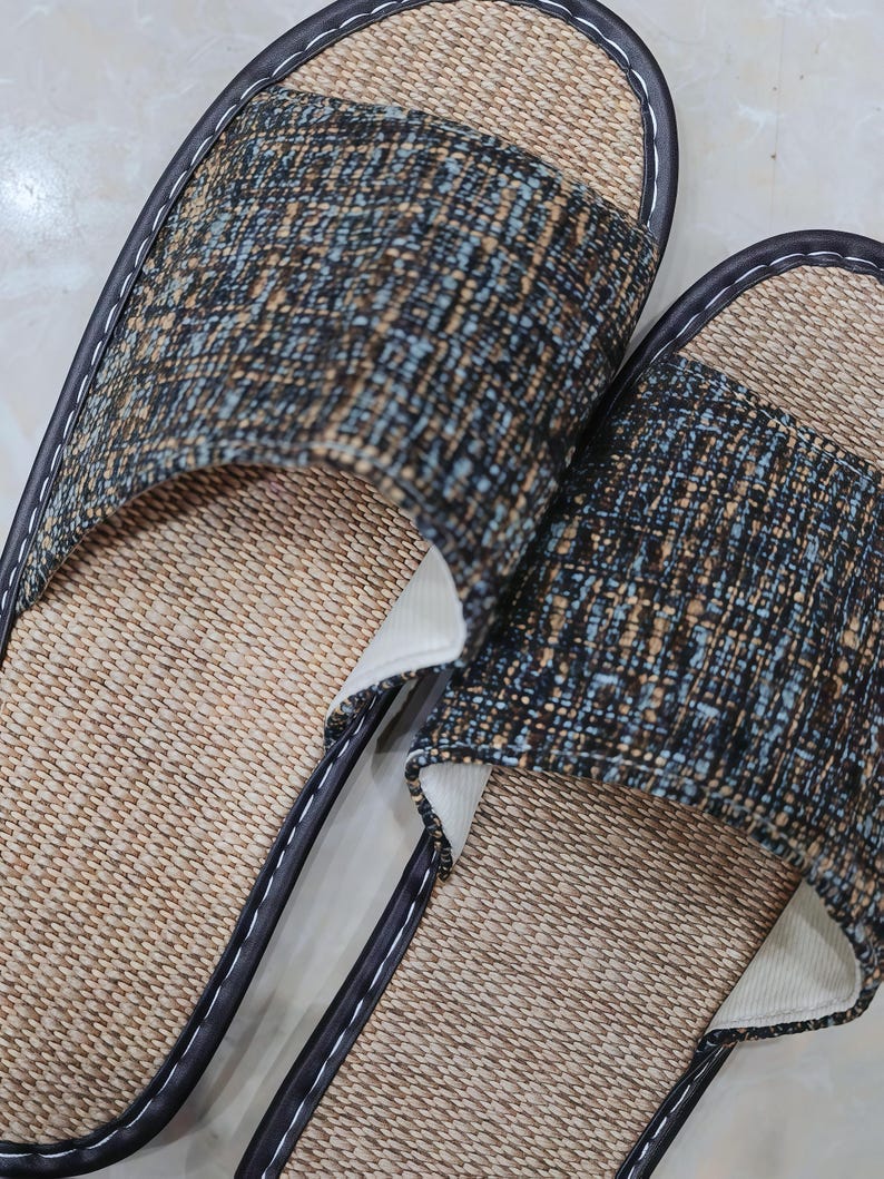 May include: A pair of open-toe slippers featuring a woven tan straw-like footbed and a wide upper strap made of a textured fabric in shades of dark blue, brown, and light blue. The edges are finished with dark brown piping and white stitching.