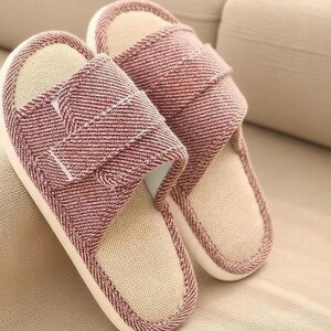 May include: A pair of red and white striped fabric slippers. The slippers have a beige interior and a white sole. The top strap is adjustable with a hook and loop closure. The slippers are resting on a beige surface.