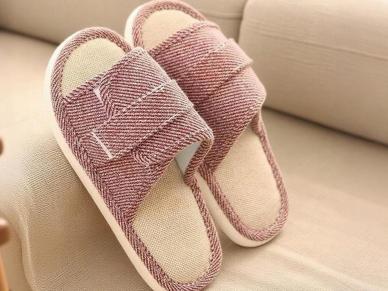 May include: A pair of red and white striped fabric slippers. The slippers have a beige interior and a white sole. The top strap is adjustable with a hook and loop closure. The slippers are resting on a beige surface.