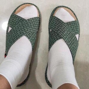 K&ouml;nnte beinhalten: Ein Paar gr&uuml;n gewebte Sandalen mit Kreuzmuster, getragen mit wei&szlig;en Socken. Die Sandalen haben eine naturfarbene Einfassung und einen schlichten, l&auml;ssigen Stil, perfekt f&uuml;r den Innen- oder Au&szlig;enbereich.